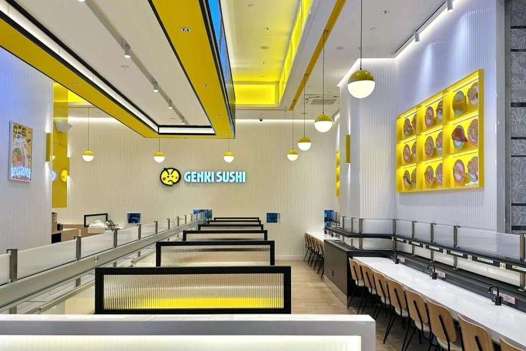 Genki Sushi features a vibrant, modern interior defined by a bold yellow and white color scheme with rows of private booths and counter seating. The restaurant is equipped with a multi-level delivery track system and decorated with circular pendant lights and a wall-mounted display of sushi plates.