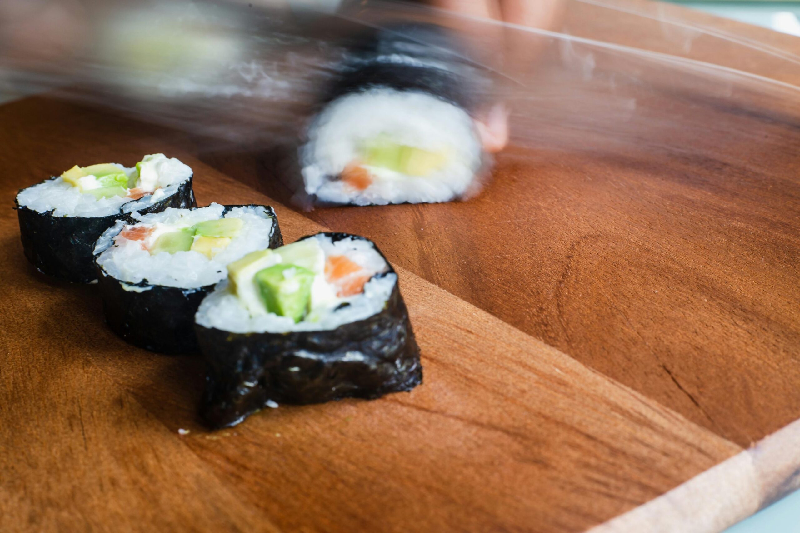  This image shows several pieces of sushi filled with avocado and salmon resting on a wooden cutting board. A blurred motion in the background suggests a knife is in the process of slicing another section of the roll.