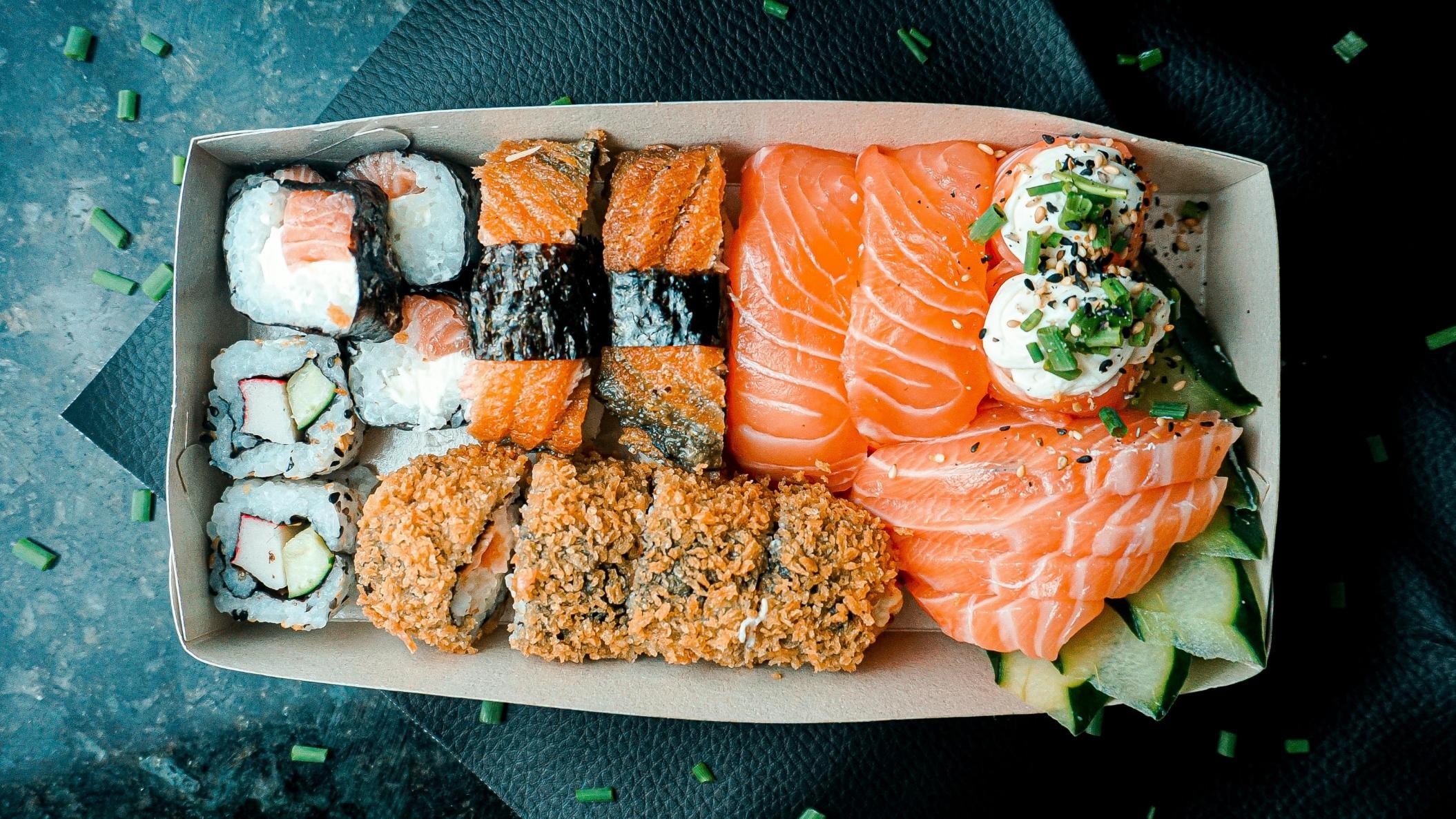An assortment of salmon sushi, including nigiri, maki rolls, and crispy fried pieces, is neatly arranged in a cardboard takeout container and garnished with fresh scallions and sesame seeds.