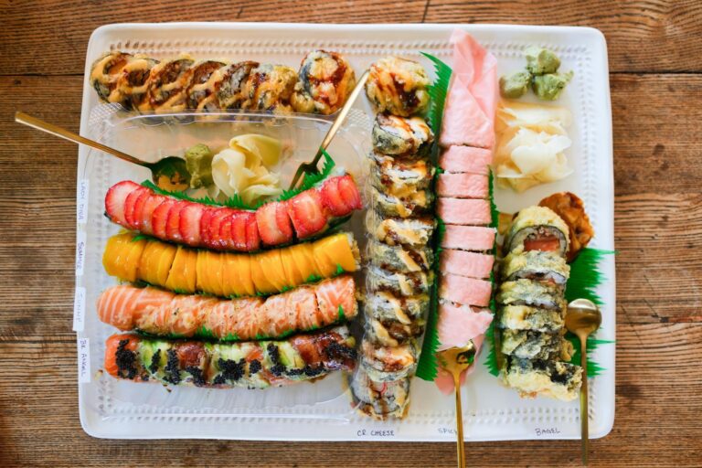 An overhead shot displays an assortment of colorful sushi rolls, including fruit-topped varieties and fried tempura rolls, meticulously arranged on a large white platter. Small handwritten labels identify the different rolls, while garnishes of pickled ginger and wasabi sit alongside elegant gold utensils.