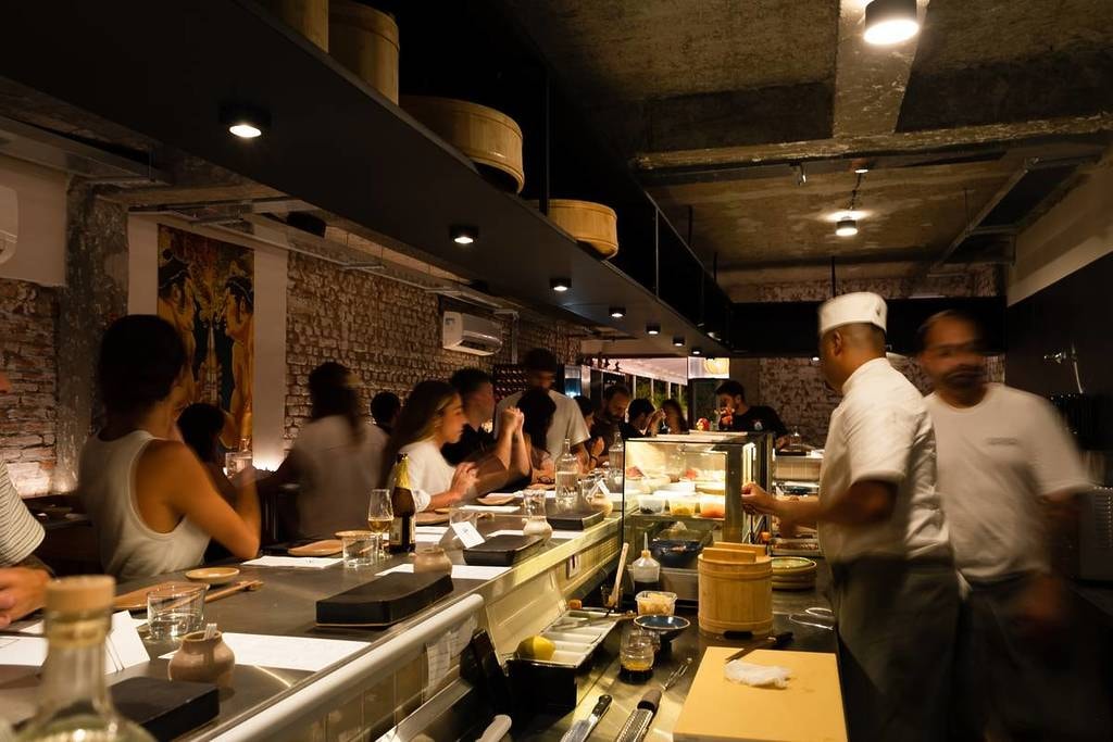 A bustling sushi bar features chefs in white uniforms preparing food behind a long counter for a line of seated customers. The restaurant's industrial-chic aesthetic is defined by exposed brick walls, dim spotlighting, and wooden storage barrels tucked into overhead shelving.