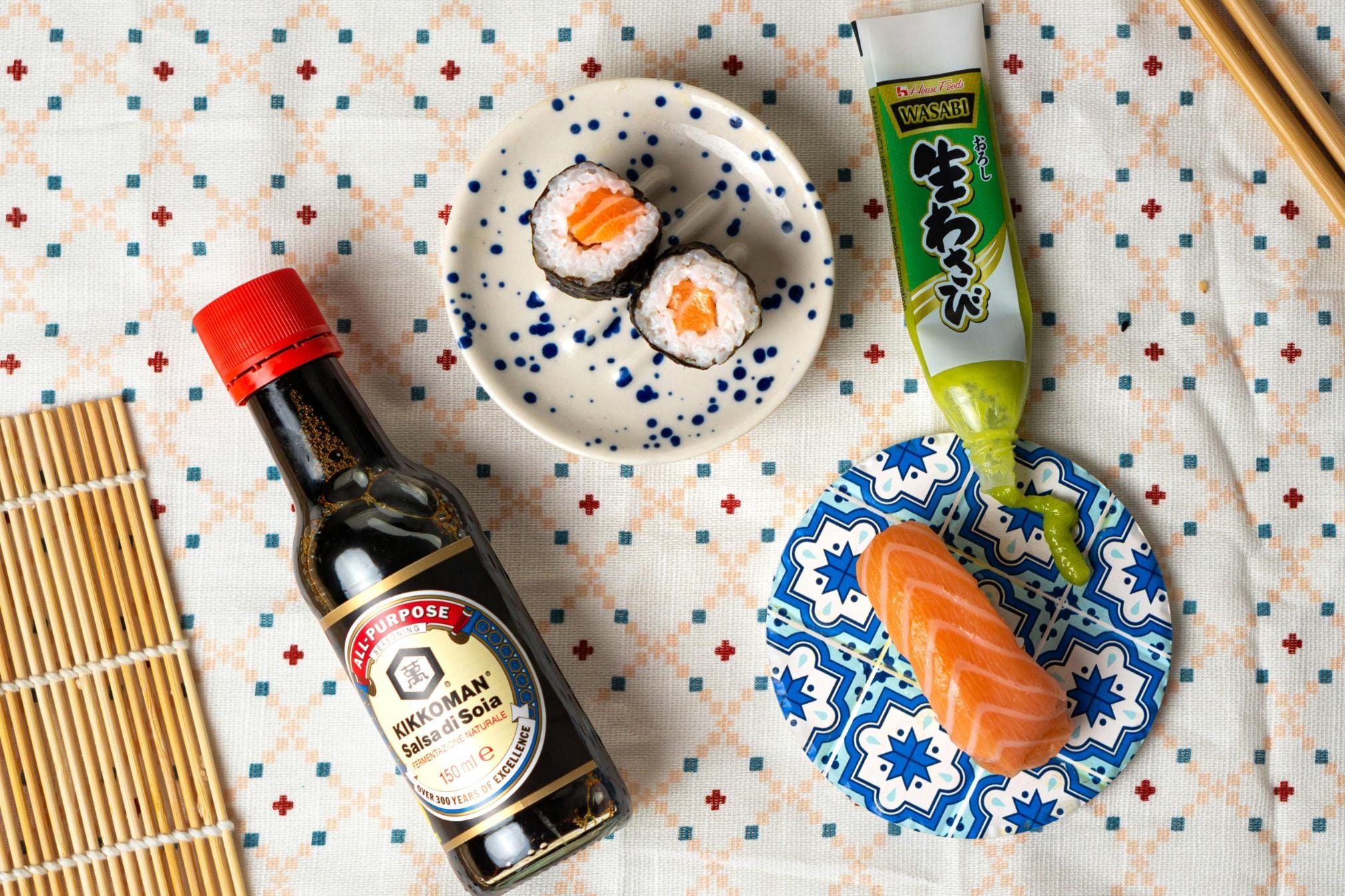 A top-down view shows fresh salmon sushi and maki rolls arranged on patterned ceramic plates alongside a bottle of Kikkoman soy sauce. A tube of wasabi paste is being squeezed onto the surface, all set against a white patterned tablecloth with a bamboo rolling mat and chopsticks nearby.