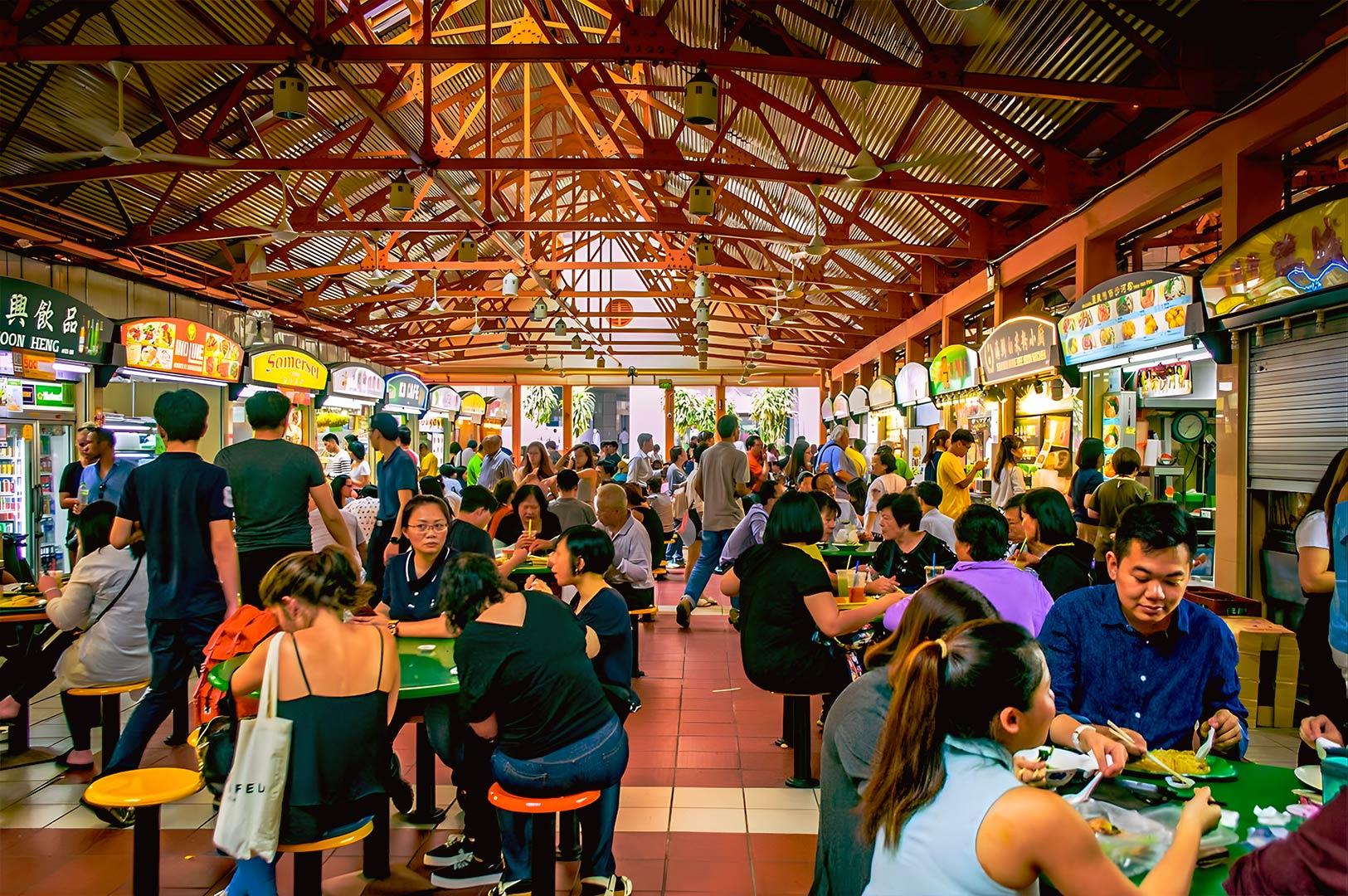 Crowded Singapore hawker center with diverse food stalls and diners, representing multicultural dining culture and the rise of affordable sushi in hawker settings