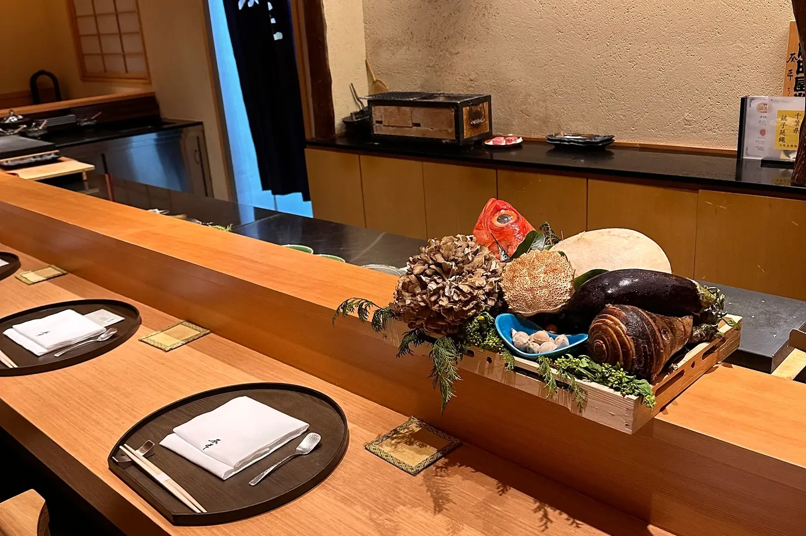A counter with place settings in a restaurant, featuring a display of vegetables, mushrooms, and seafood on a wooden tray.