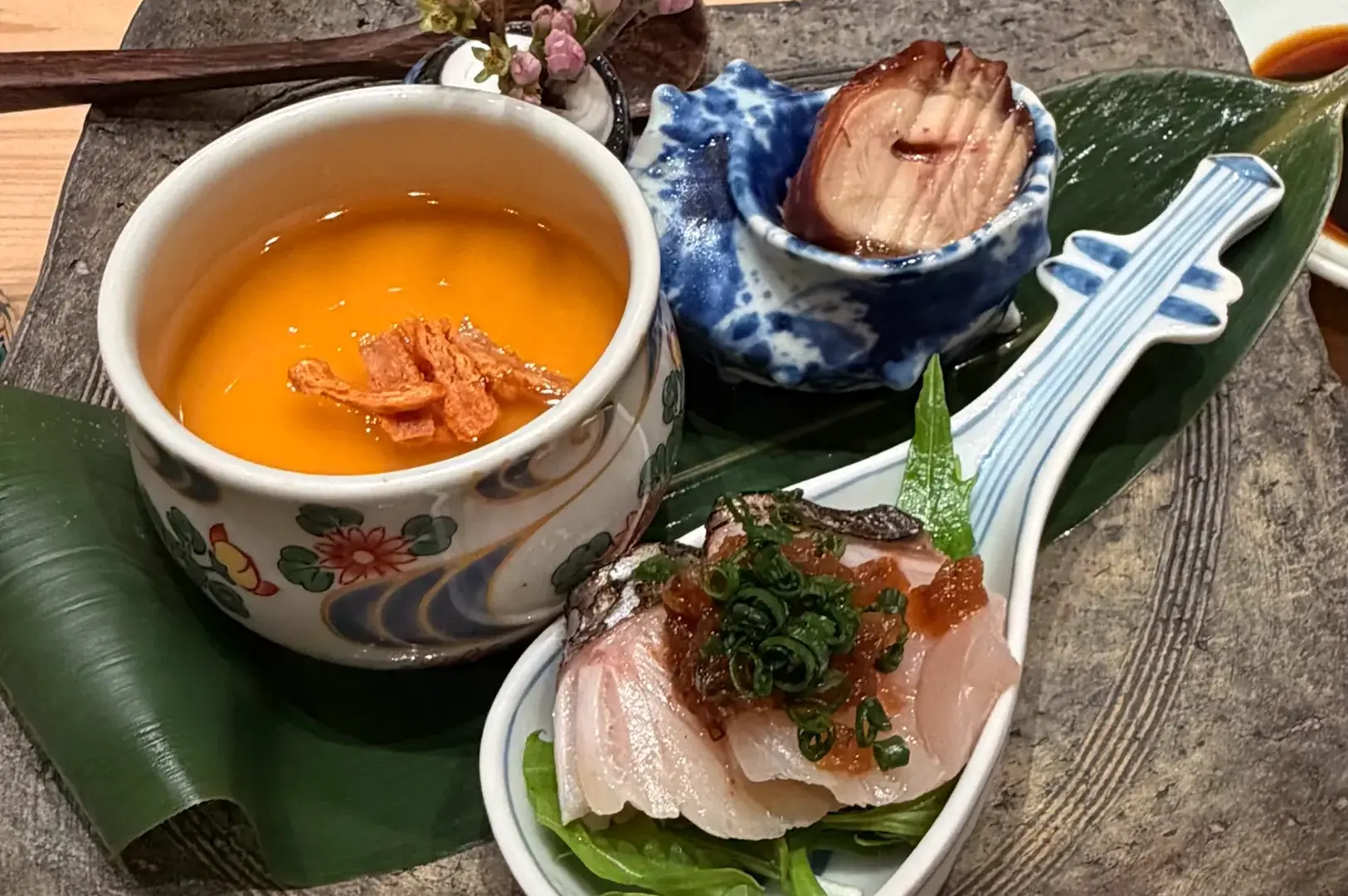 Elegant omakase presentation on stone plate featuring golden soup in decorative bowl, fresh sashimi with garnish on white dish, and green leaf accent in bright editorial lighting