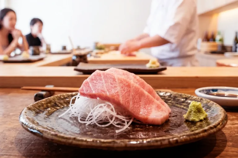 Premium sashimi slice on dark ceramic plate with shredded daikon and wasabi in foreground, omakase chef in white uniform