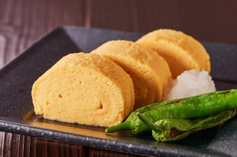 A close-up shot of sliced tamagoyaki, a Japanese rolled omelet, served with grated daikon radish and grilled shishito peppers on a dark rectangular plate.