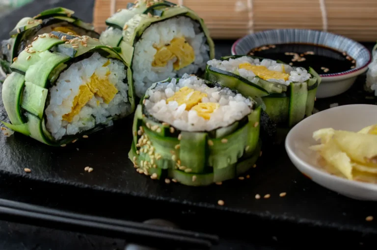 Several pieces of sushi rolled in cucumber strips and sprinkled with sesame seeds, served with soy sauce and pickled ginger.