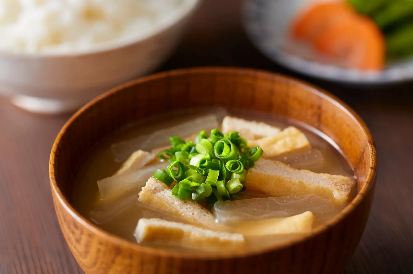 A bowl of miso soup with tofu and green onions, served with rice and pickled vegetables.