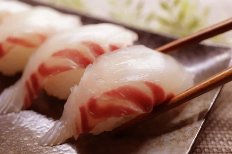 Close-up of nigiri sushi with white fish and red stripes on a dark plate, held by chopsticks.