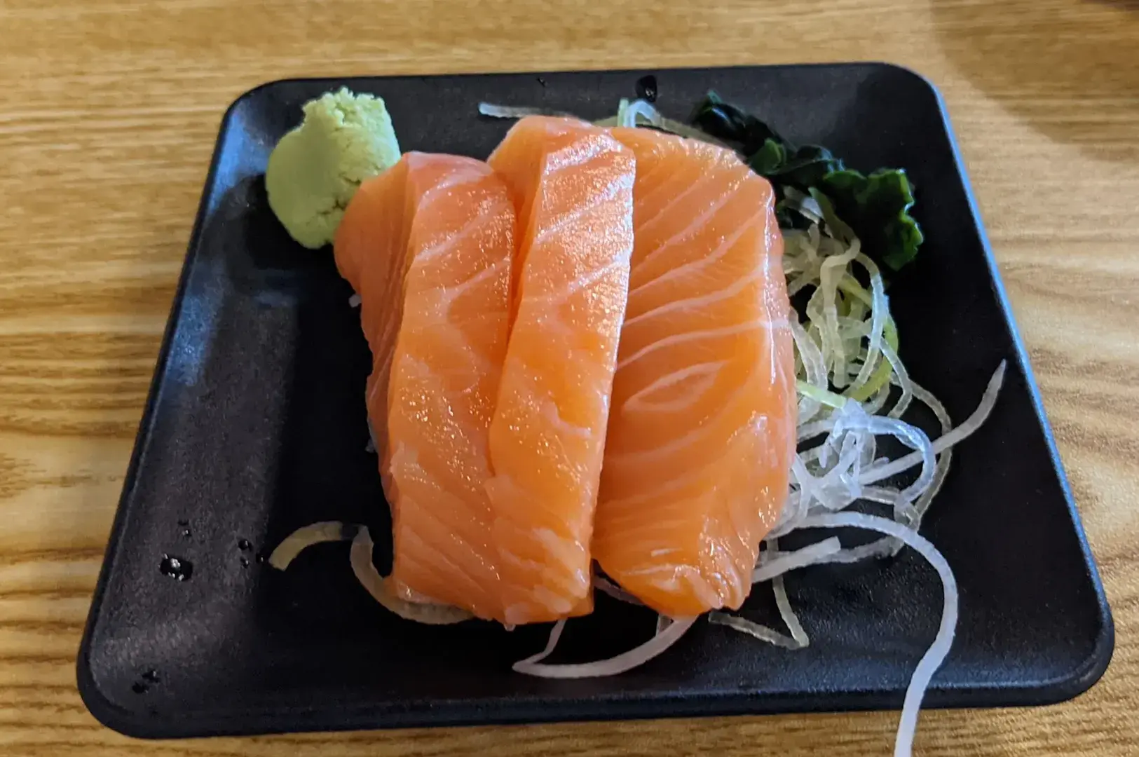 A black plate of salmon sashimi with wasabi and shredded garnish on a wooden table.