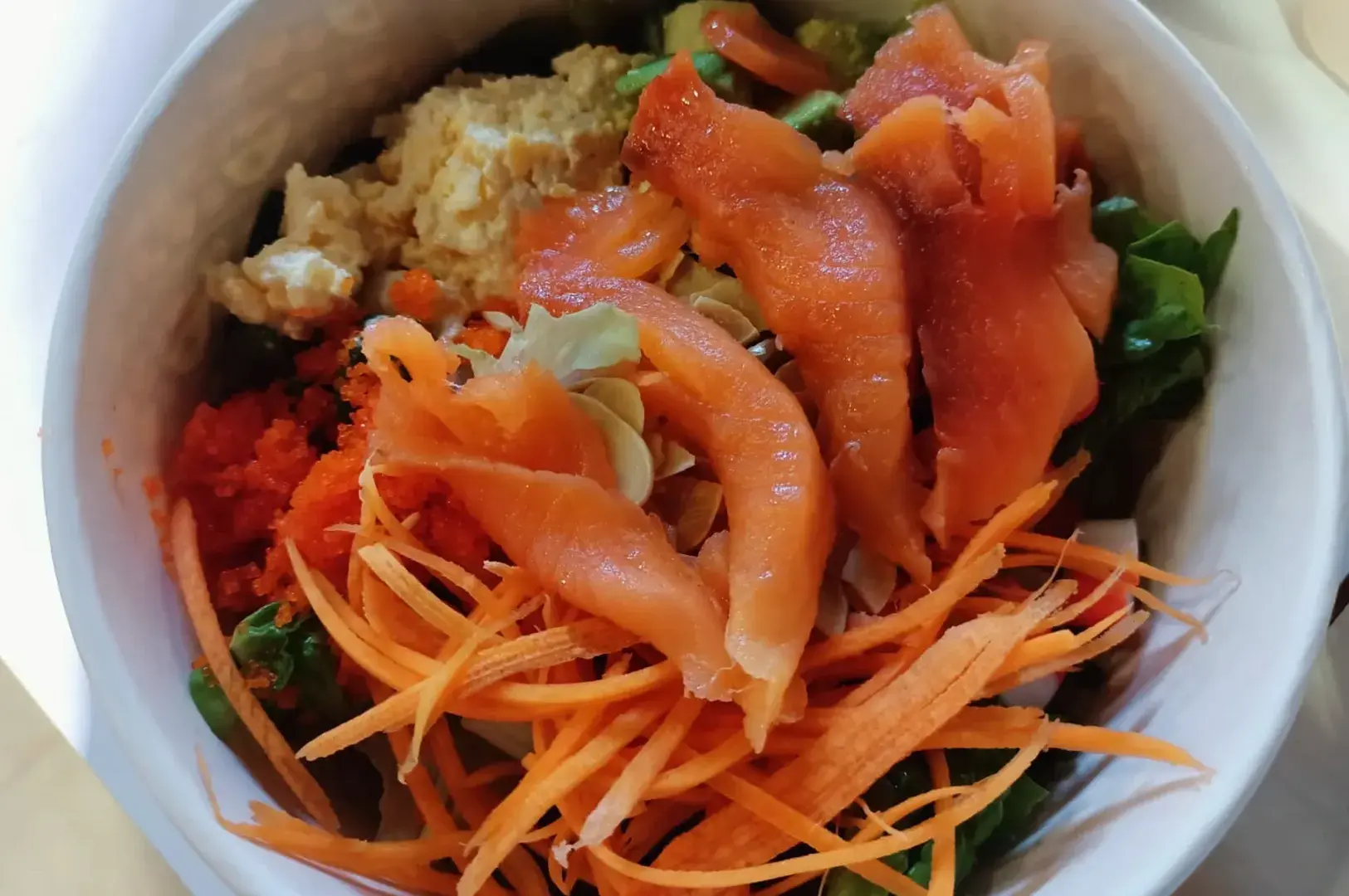 A sushi bowl topped with smoked salmon, shredded carrot, greens, and sauces.