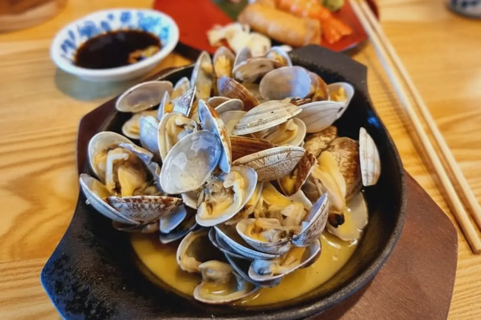 A black skillet of clams in sauce with chopsticks and a dipping dish on a wooden table.