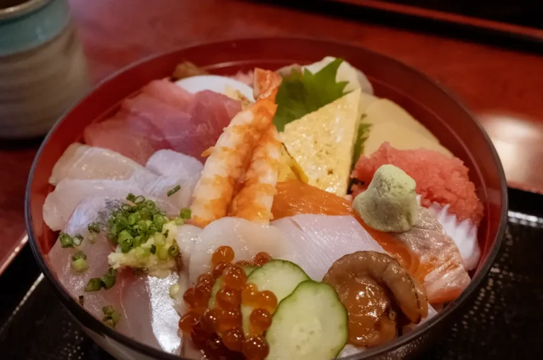 Chirashi bowl with assorted sashimi including ebi, ikura, tamago, salmon, tuna, and wasabi in red lacquer bowl.