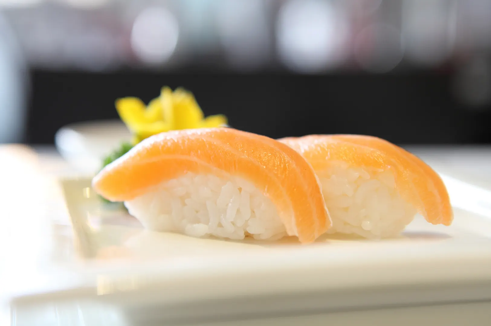 Two pieces of salmon nigiri sushi on a white plate with pickled ginger in the background.