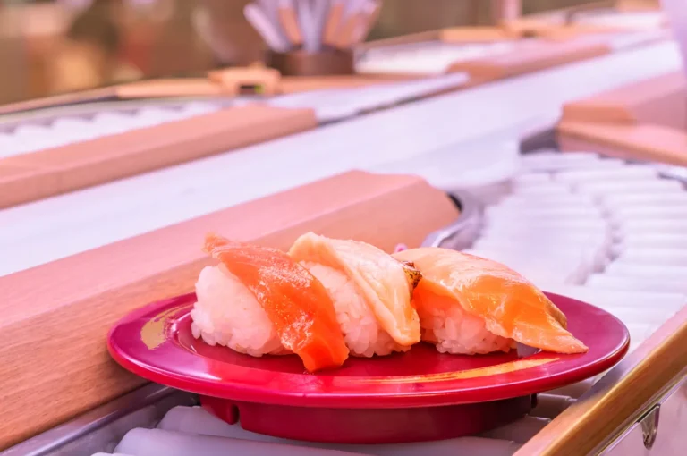 A red plate with three pieces of sushi on a conveyor belt at a sushi restaurant.
