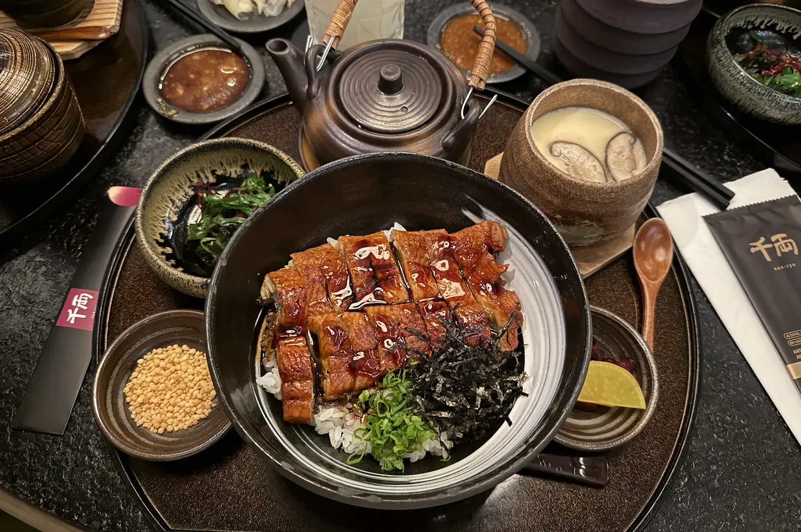 Unagi donburi with rice, nori, and scallions on dark table setting.