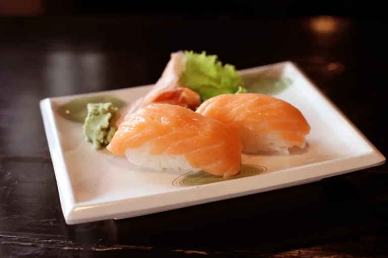 Two slices of salmon nigiri on a white rectangular plate, topped with green garnish and a small mound of wasabi, set on a dark table.