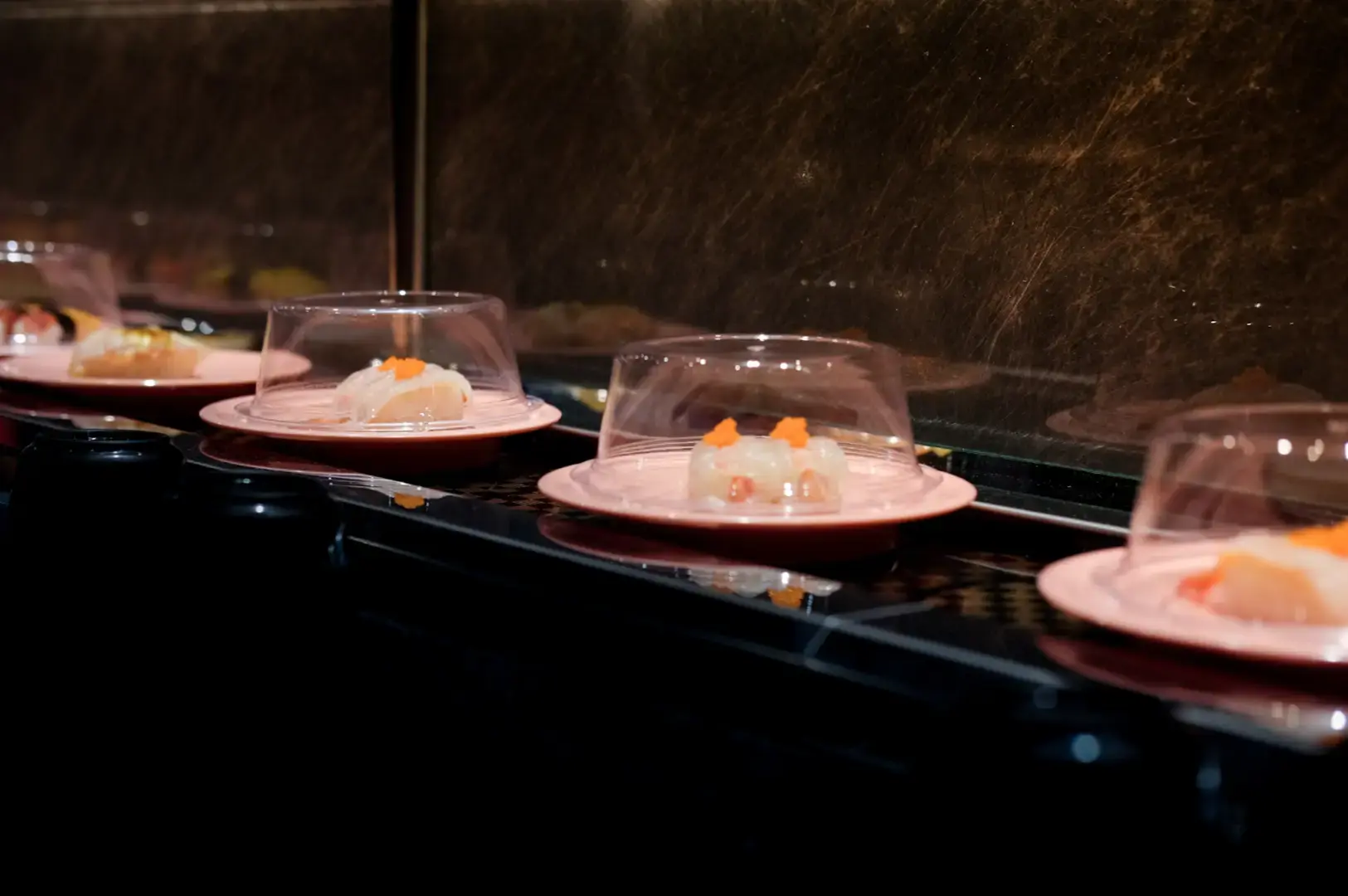 Row of clear plastic food covers on a dark counter, each holding a small prepared dish with visible orange and pink toppings.