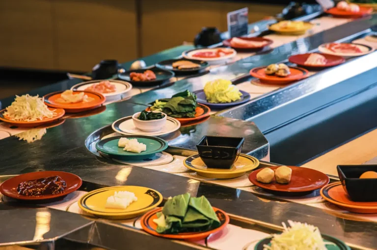 A sushi belt station with rows of small plates and bowls containing sliced meats, vegetables, sauces, and garnishes arranged along a long counter.