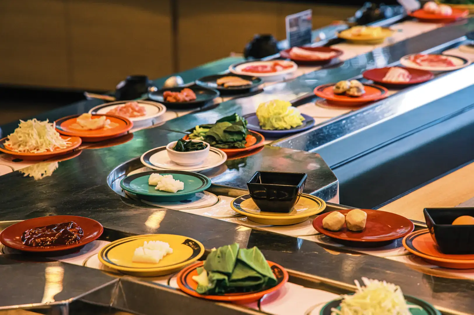 A sushi belt station with rows of small plates and bowls containing sliced meats, vegetables, sauces, and garnishes arranged along a long counter.