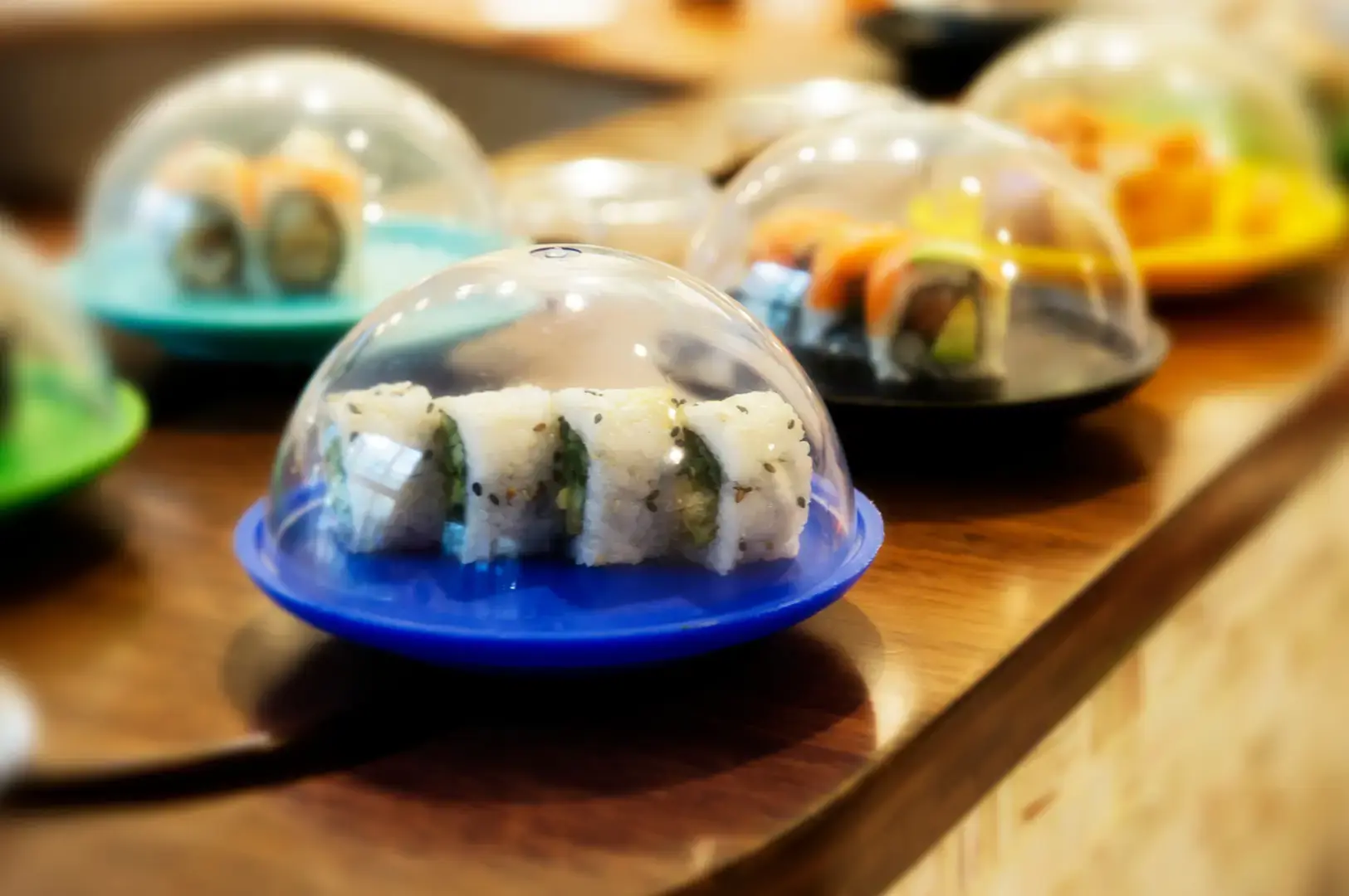 A blue plastic dome on a wooden table covering a small dishes with white and green layers, with other domed treats blurred in the background.