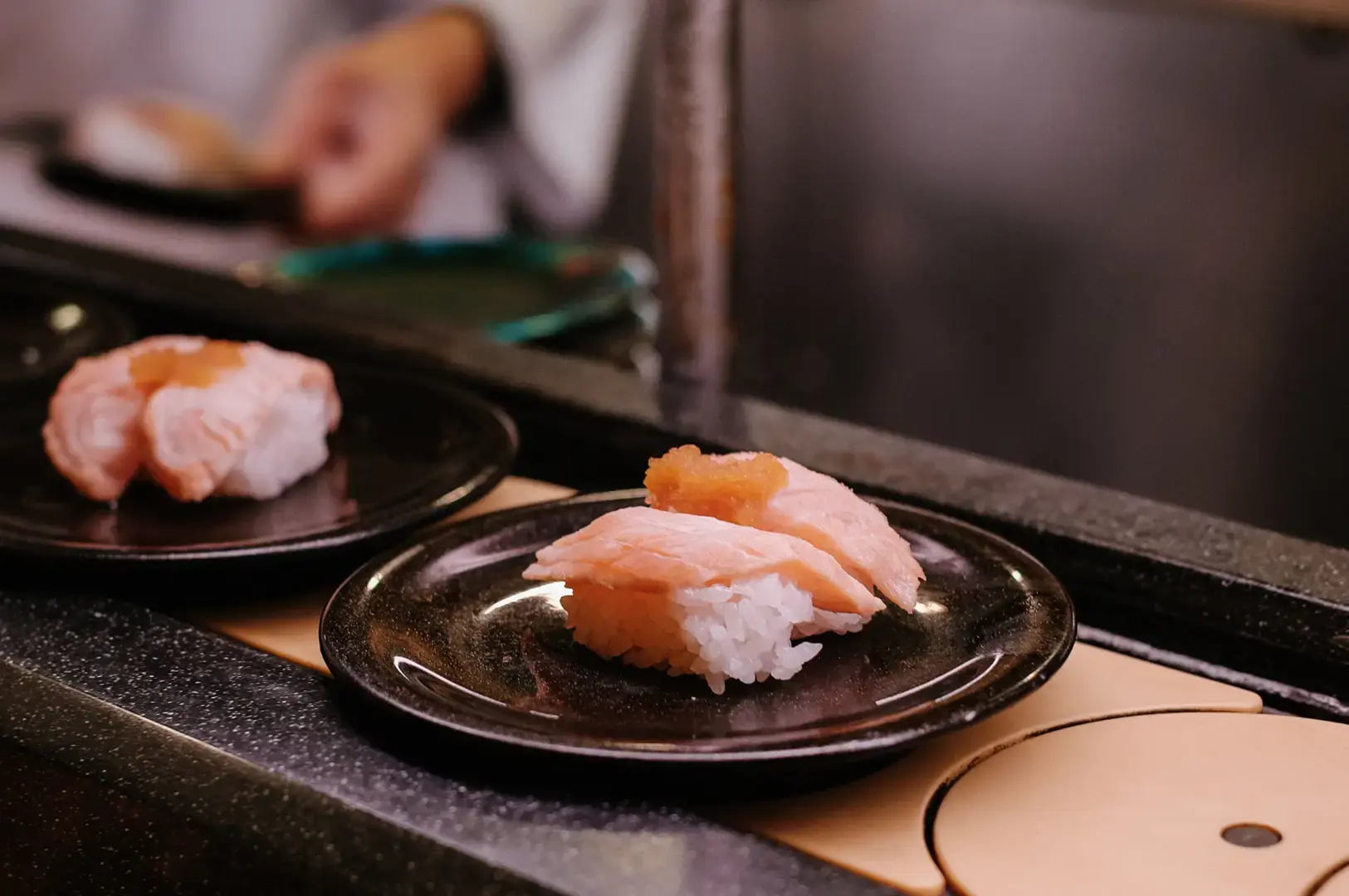 Several pieces of sushi with orange topping sit on small black plates on a conveyor-style kitchen counter, with a blurred worker in the background.