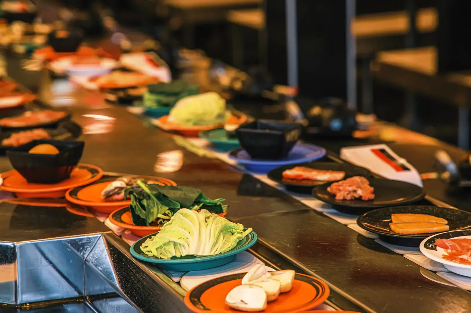 A sushi belt station with multiple black plates and orange-rimmed dishes lined up on a metal counter, holding sliced mushrooms, cabbage, and pieces of meat, with blurred items and warm lighting in the background.