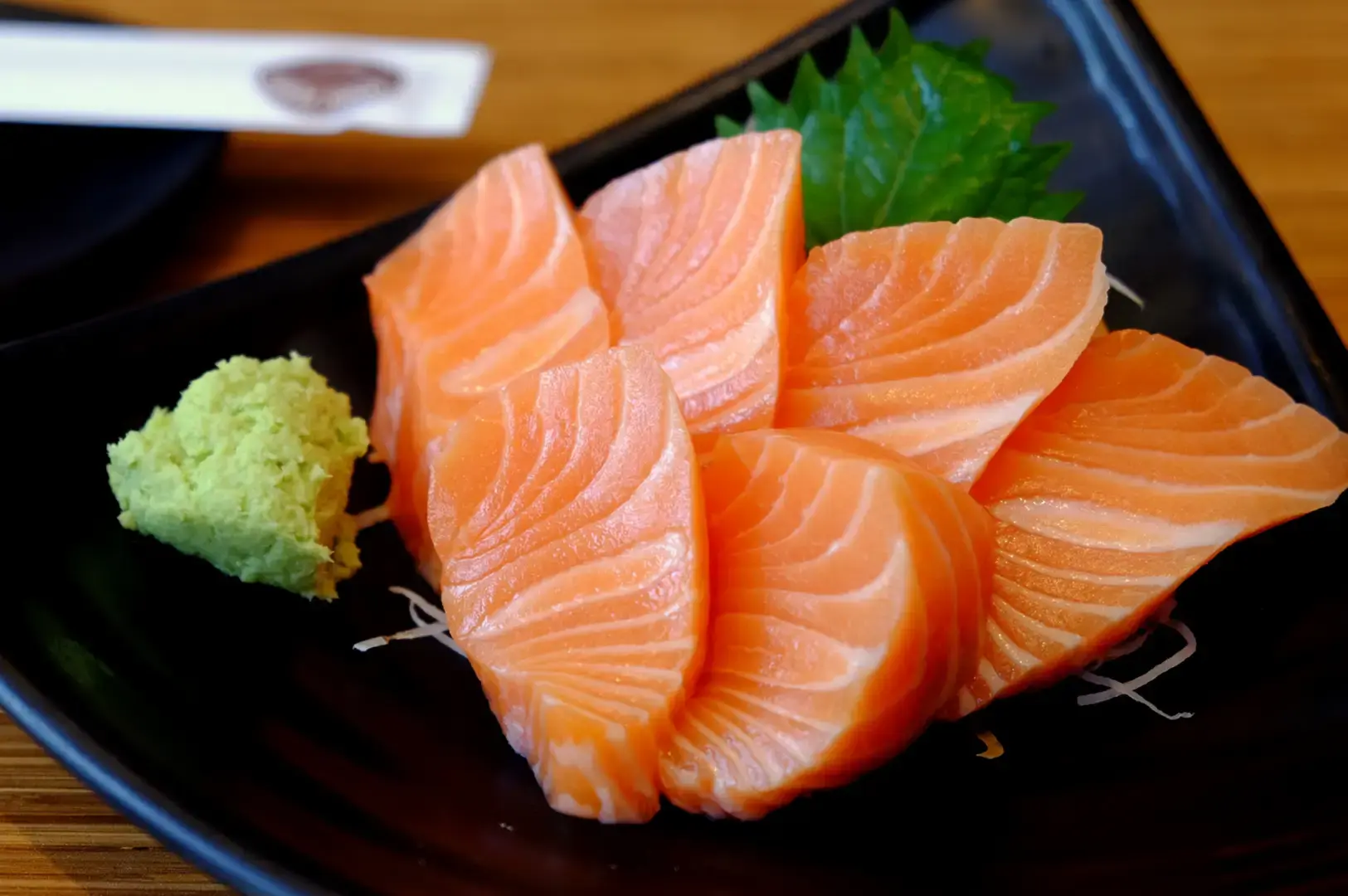 Thick slices of fresh salmon sashimi arranged on a black plate with shiso leaves, shredded daikon, and a mound of wasabi.