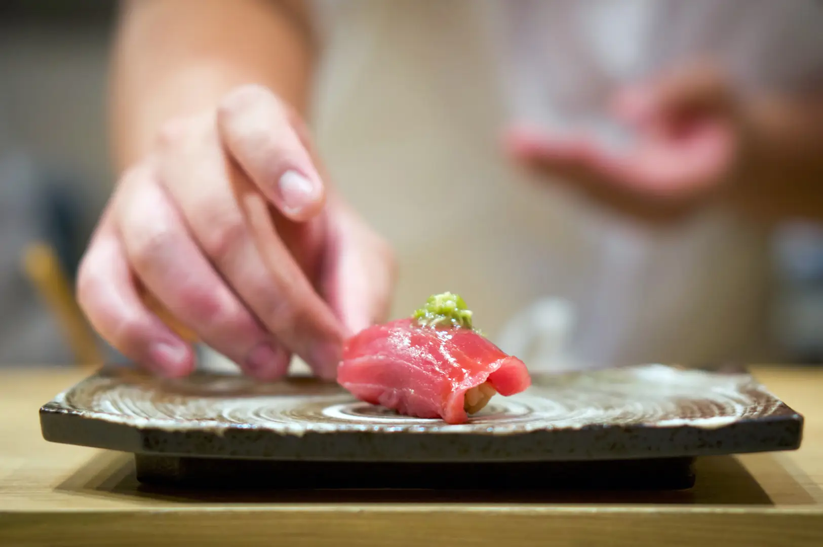 Hands slice a small piece of red tuna with a knife on a dark wooden chopping board, with another hand reaching in the blurred background.