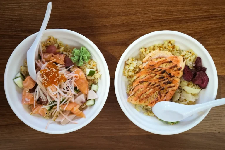 Two bowls of rice topped with salmon, vegetables, pickled sides, and white spoons on a wooden table.