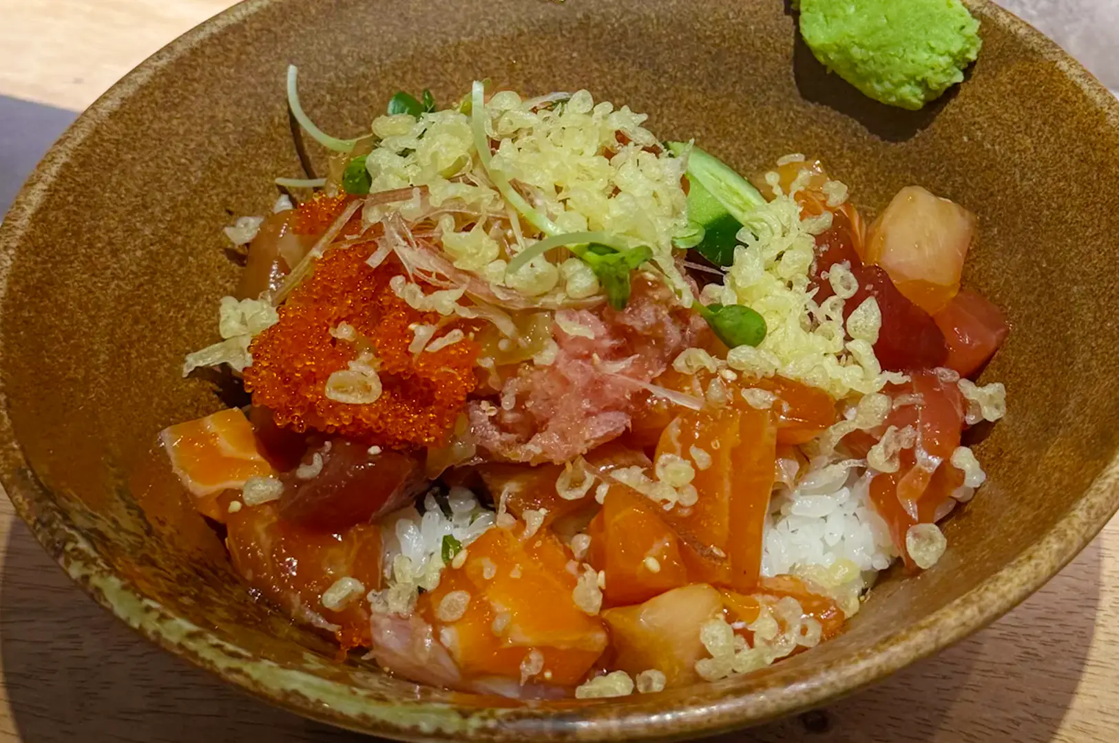 Bowl of chirashi sushi rice topped with diced salmon, tuna, orange fish roe, spring onions, tempura flakes, and wasabi.