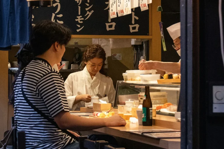 A customer sits at a sushi counter while a chef prepares food behind the display case, with Japanese menu boards, sushi ingredients, and a green bottle visible in the warm-lit restaurant.