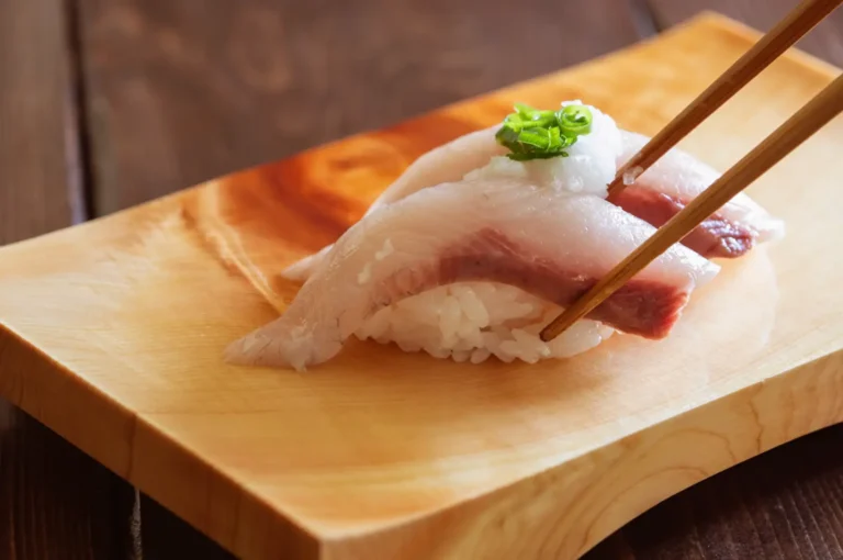 Piece of nigiri sushi on a light wooden board, topped with white fish, a strip of pink cured fish, and a small garnish of chopped green onions, held with wooden chopsticks.