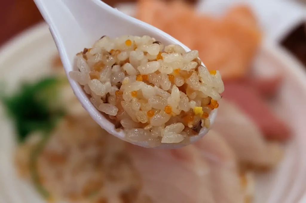 Close-up of seasoned rice with salmon roe on a white spoon, with a bowl of food blurred in the background.