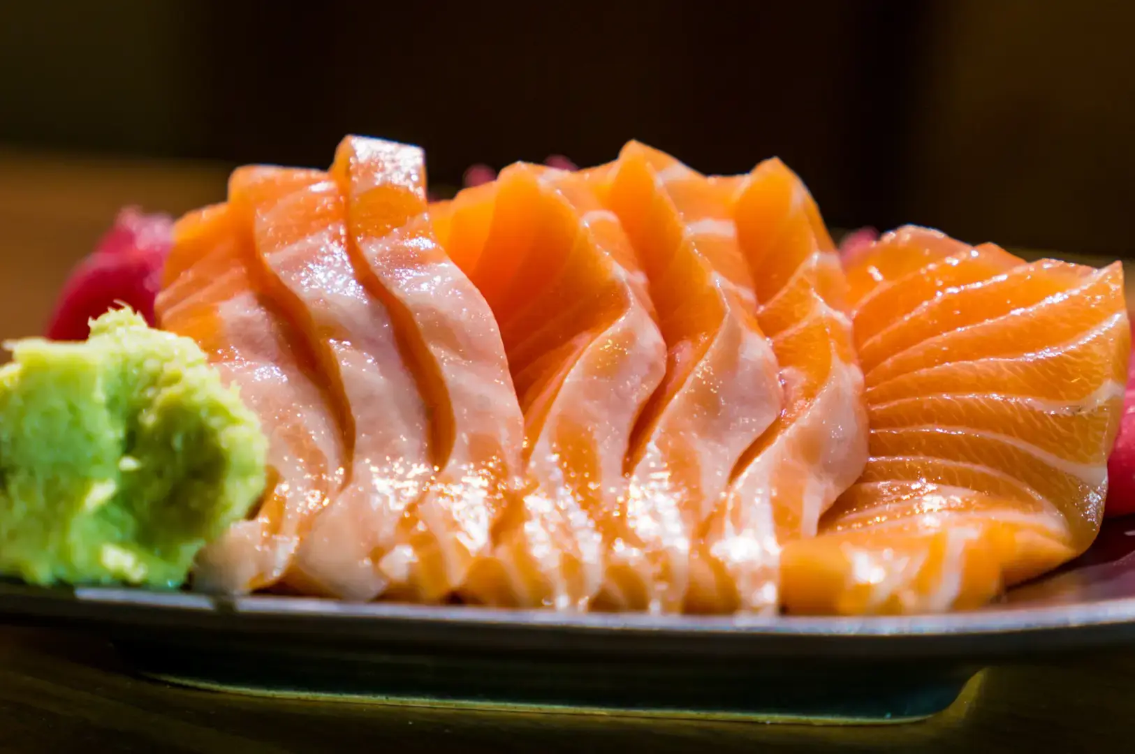 Close-up of glossy salmon sashimi slices arranged on a black plate with wasabi and shredded daikon in the background.