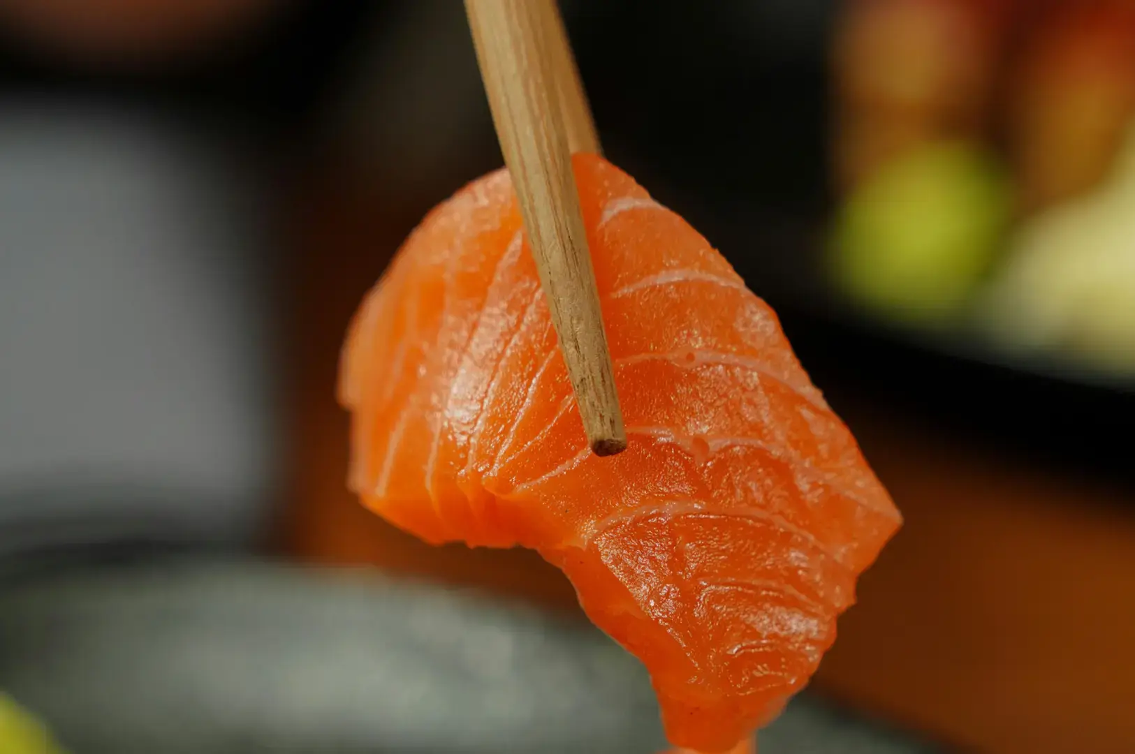 A slice of salmon sashimi held with wooden chopsticks against a softly blurred restaurant background.