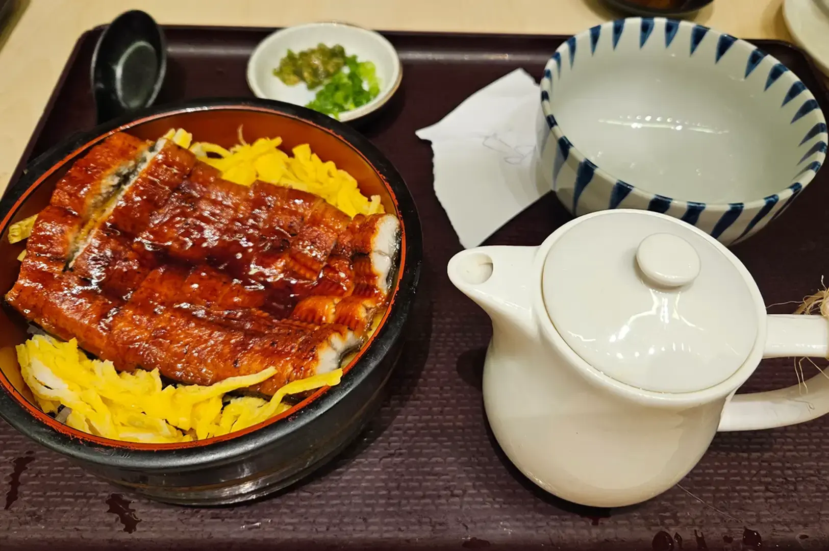 Glazed grilled eel served over yellow rice in a black bowl beside a white teapot, dipping dish, spoon, and condiments on a restaurant tray.