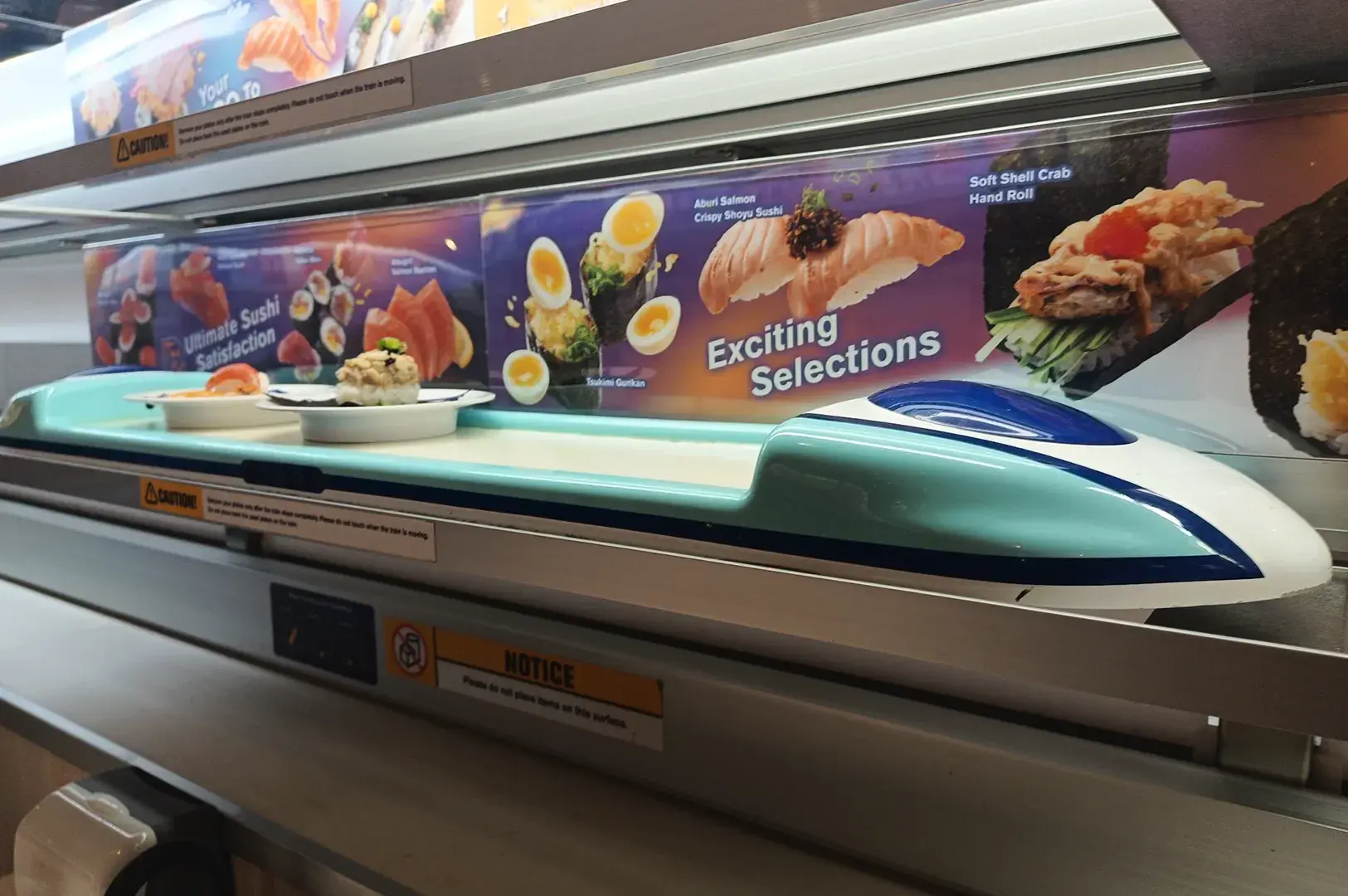 Sushi-Go conveyor belt carrying sushi plates beneath a display advertising assorted sushi selections.