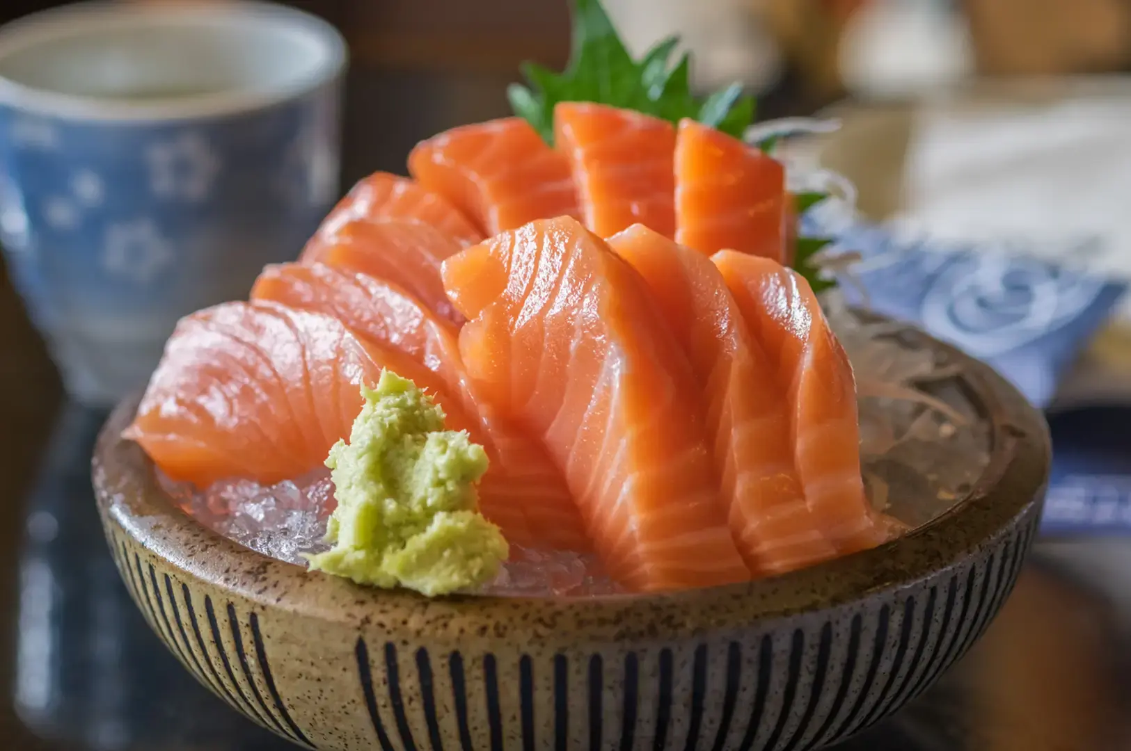 Bowl of salmon sashimi served over crushed ice with wasabi and shiso leaves, accompanied by a cup and Japanese tableware.