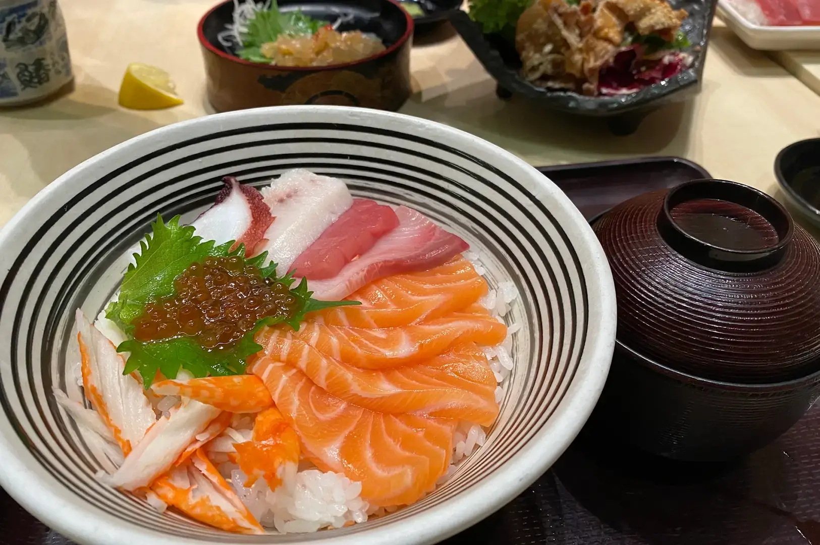 Japanese sashimi rice bowl topped with salmon, tuna, octopus, crab, and salmon roe, served with a black soup bowl and soy sauce container.
