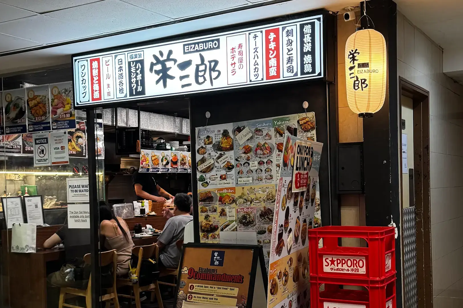 Entrance to Sakedokoro Eizaburo with illuminated Japanese signage, menu boards, a paper lantern, indoor diners, and red beverage crates.