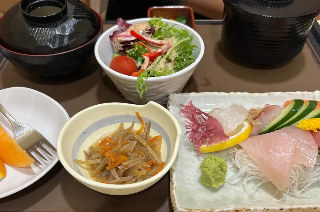 Japanese meal spread featuring assorted sashimi with wasabi and garnishes, a green salad, simmered vegetables, miso soup, rice, and side dishes arranged on a restaurant table.