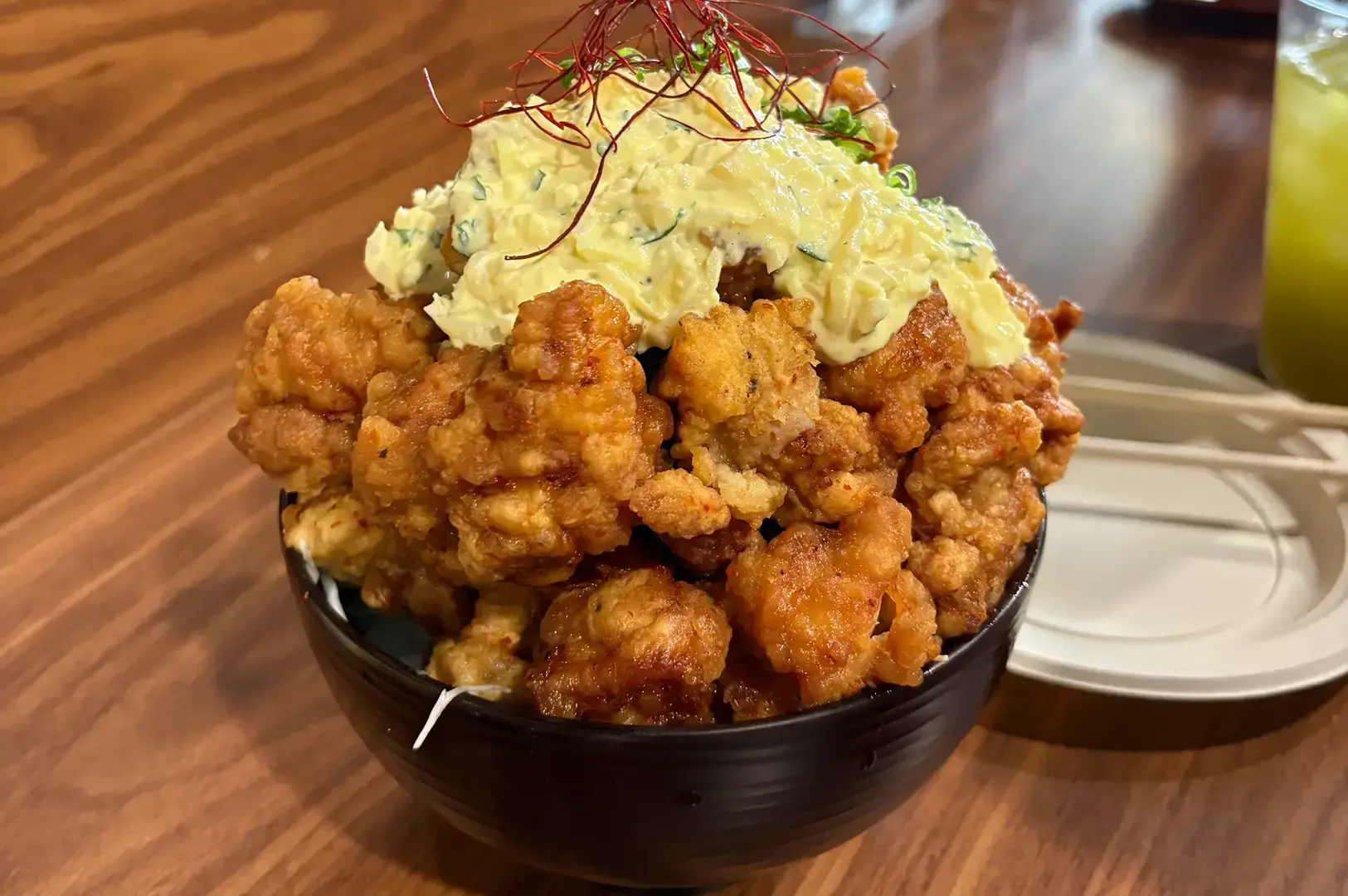 Bowl of golden fried chicken pieces topped with creamy tartar sauce and fine red chilli threads on a wooden restaurant table.