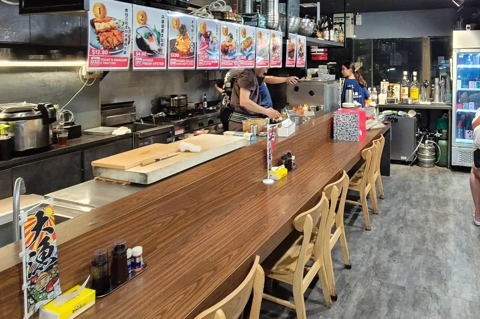 Interior of Sakedokoro Eizaburo showing a long wooden counter with stools, open kitchen, cooking equipment, menu boards, and staff preparing food.