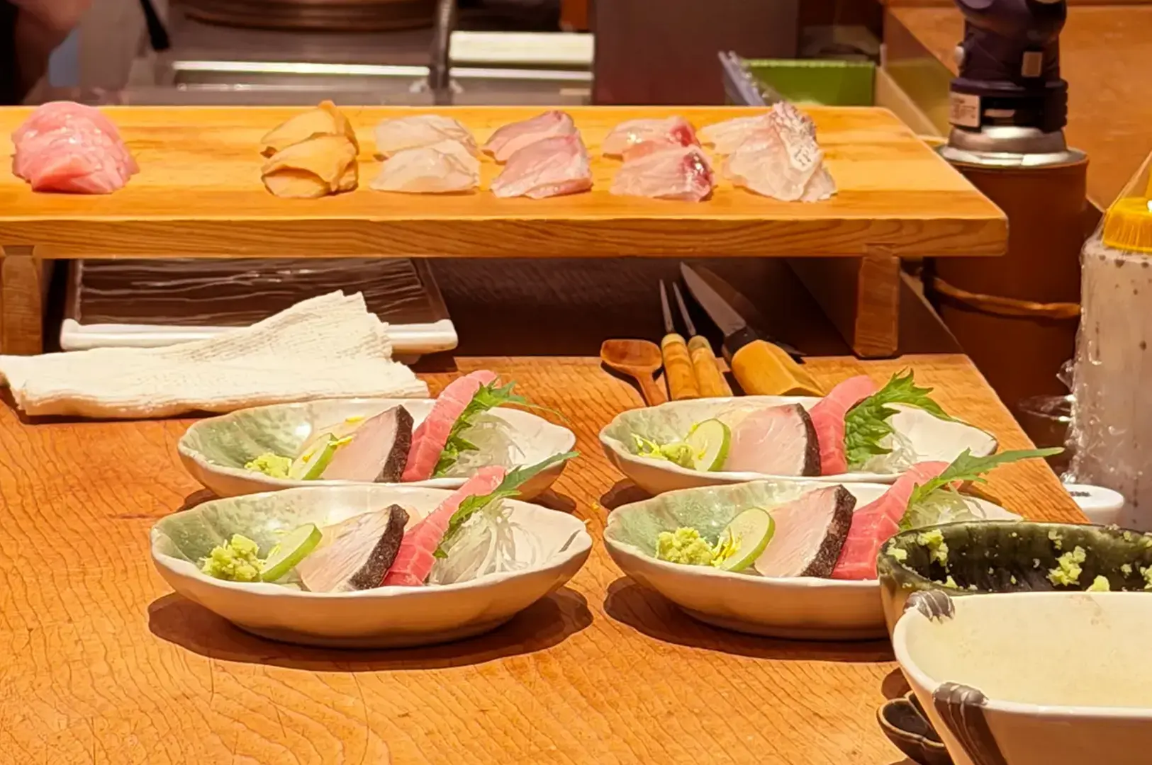 Sushi counter with assorted nigiri on a wooden board, sashimi plates garnished with shiso leaves and lime, and sushi-making tools.