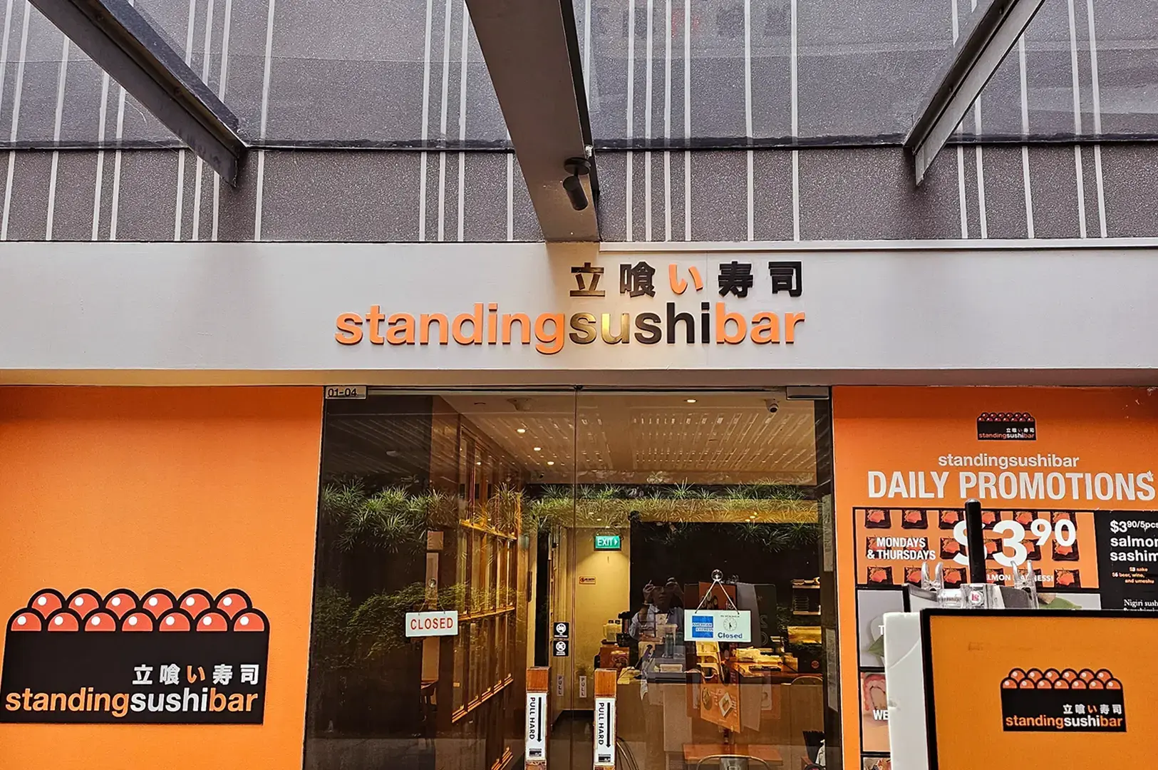 Standing Sushi Bar storefront with orange walls, glass entrance, illuminated signage, and a daily promotions board.