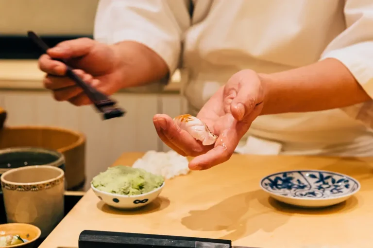 Chef preparing sushi at a wooden counter, holding a piece of nigiri in one hand while using chopsticks with the other, with wasabi, ceramic plates, a teacup, bowls, and sushi ingredients arranged across the counter.