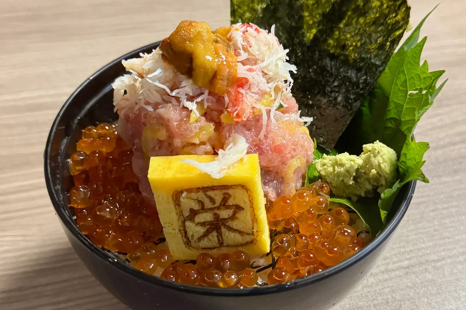 Close-up of a black bowl topped with tuna, crab, sea urchin, salmon roe, shredded seafood, egg, shiso leaves, wasabi, and roasted seaweed.