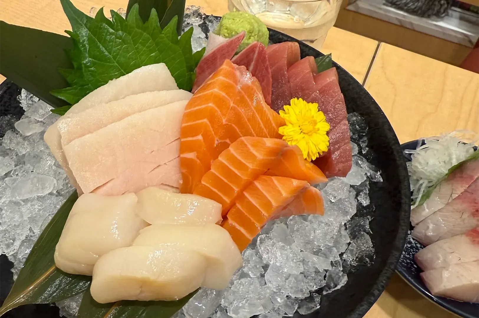 Close-up of a premium Mita Sushi sashimi platter featuring fresh scallops, salmon, and tuna served on crushed ice with edible flowers.
